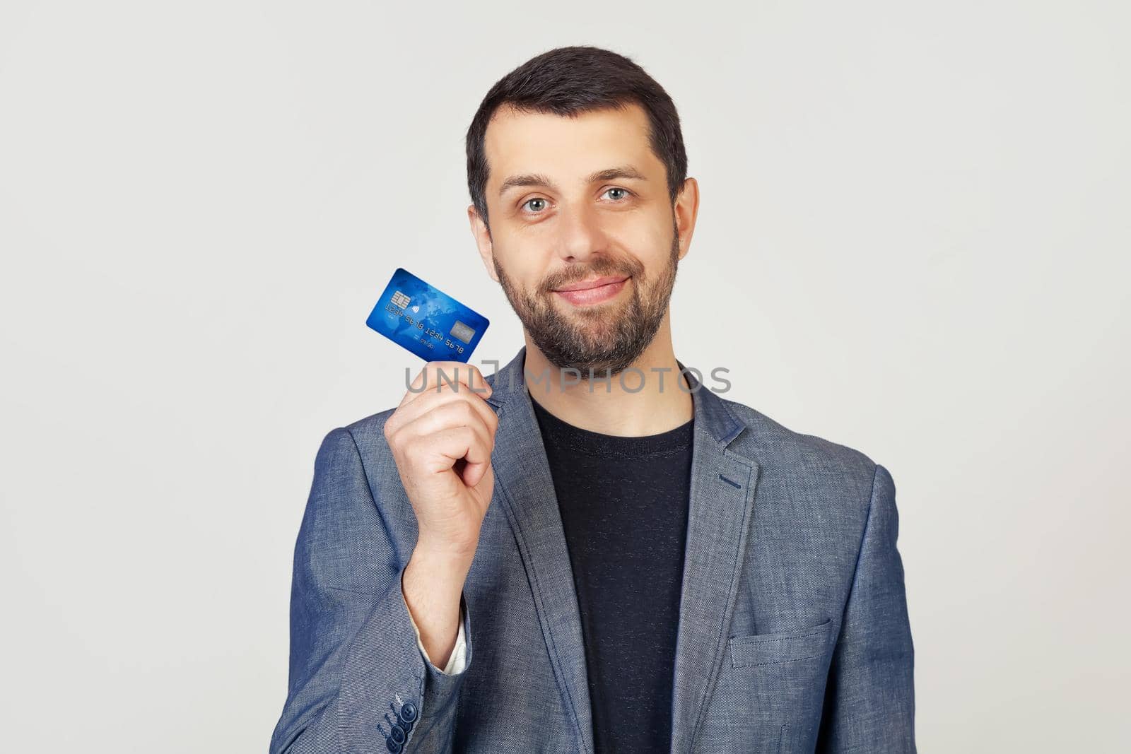 Young businessman man with a beard in a jacket holding a credit card with a confident expression on a smart face and thinking seriously. Portrait of a man on a gray background.