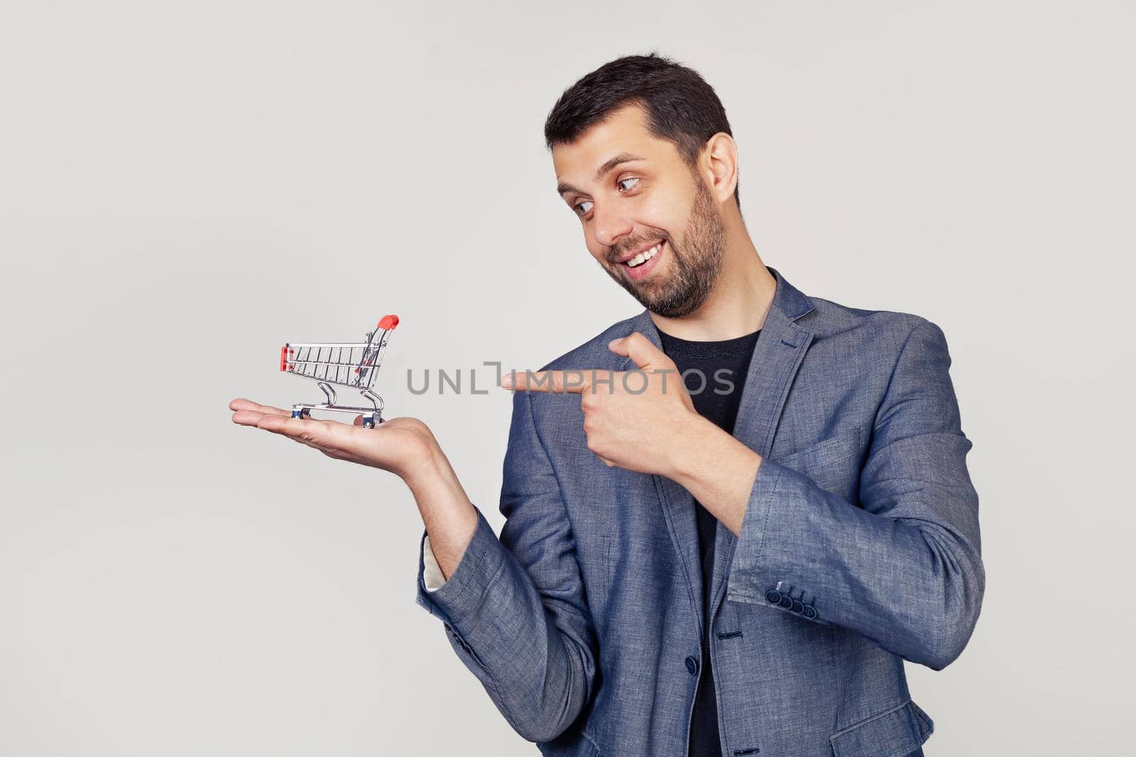 Young businessman man with a beard in a jacket looks and points to a shopping cart on his hand, showing with his hand and pointing with his finger. Portrait of a man on a gray background.