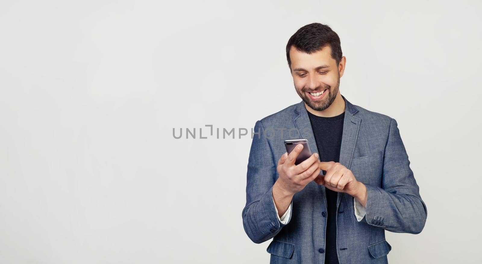 Young businessman man with a beard in a jacket, reading a message with good news on a smart phone with a happy face and smiling with a confident smile showing teeth. man on a gray background.