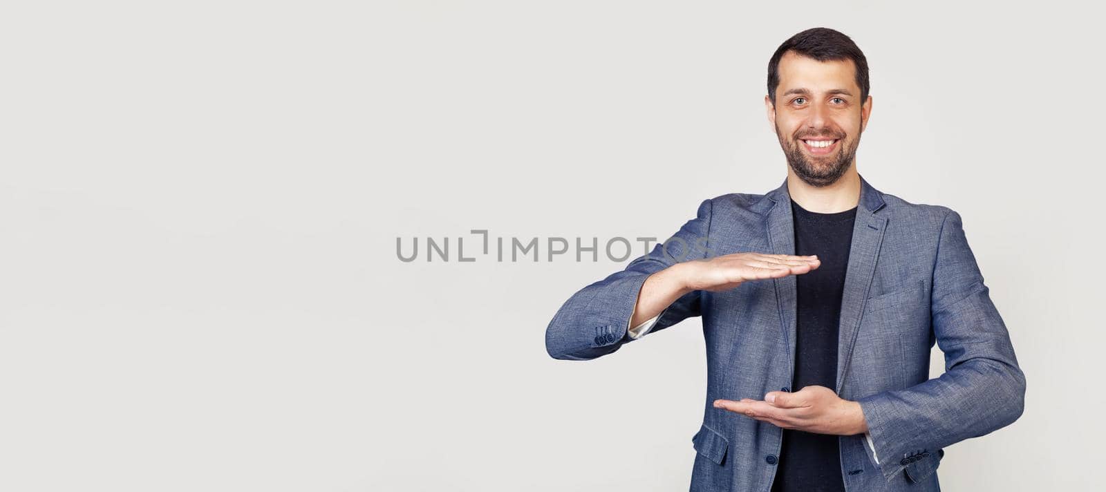 Young businessman man with a beard in a jacket, gesturing with his hands showing a large and large size sign a symbol of measure. Smiling looking into the camera Portrait of a man on a gray background by ViShark