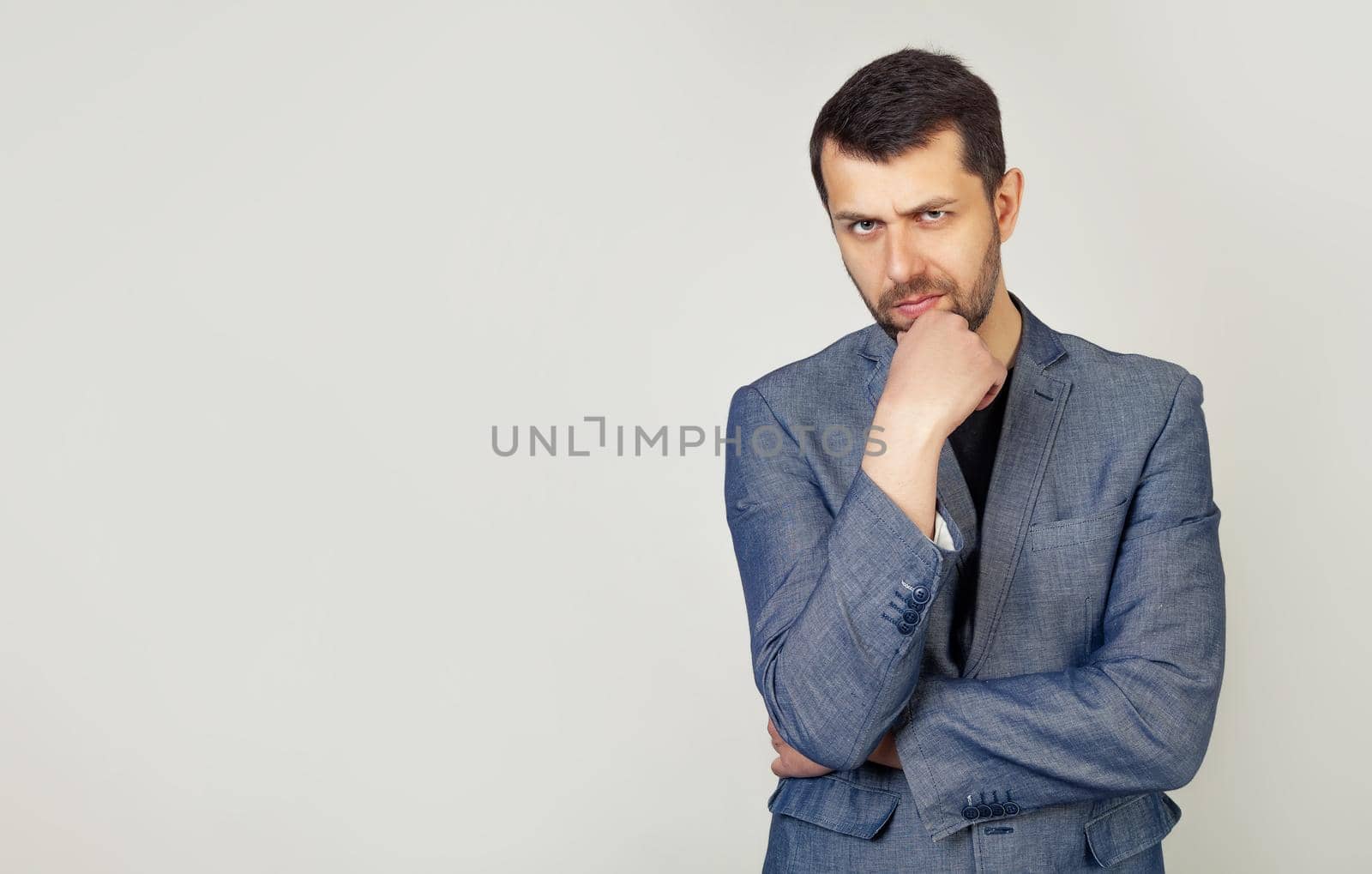 The man is looking thoughtfully at the camera. Confident young man with a beard in a stylish jacket, holds his hand on his chin, raised eyebrow thinks about something makes a plan, ponders a cool idea
