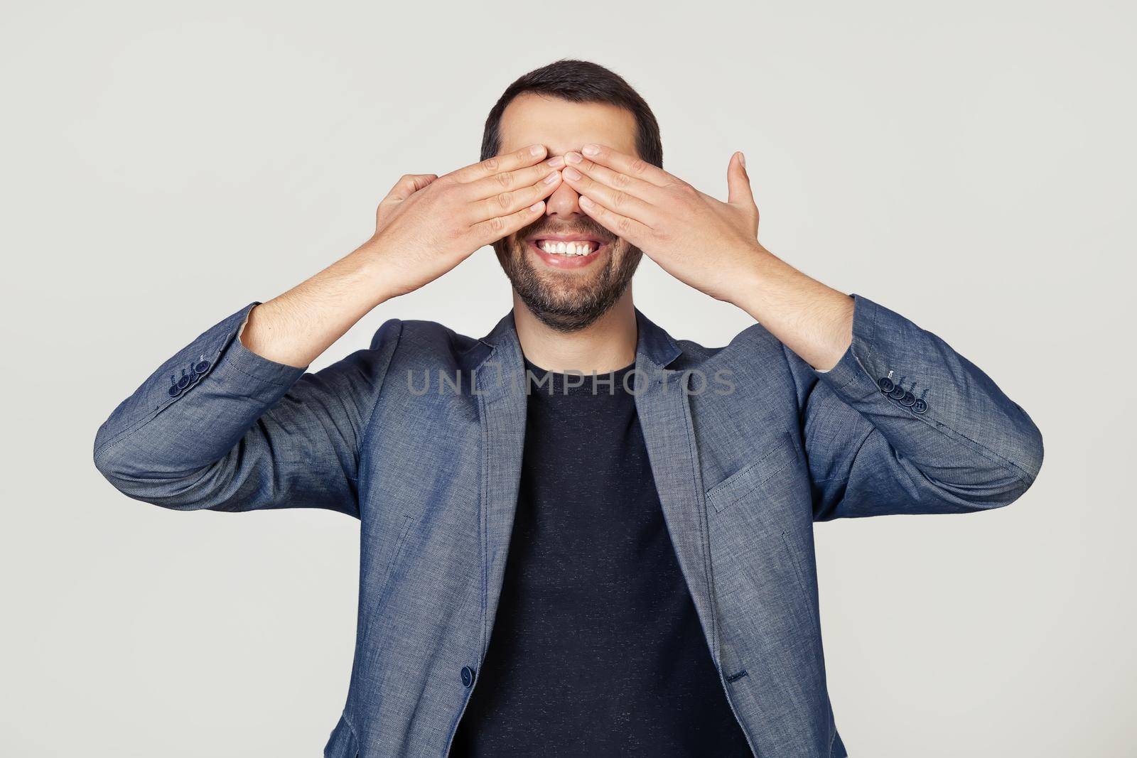 Young businessman man with a beard in a jacket covering his eyes with his hands smiling cheerful and funny. Blind concept. Portrait of a man on a gray background.
