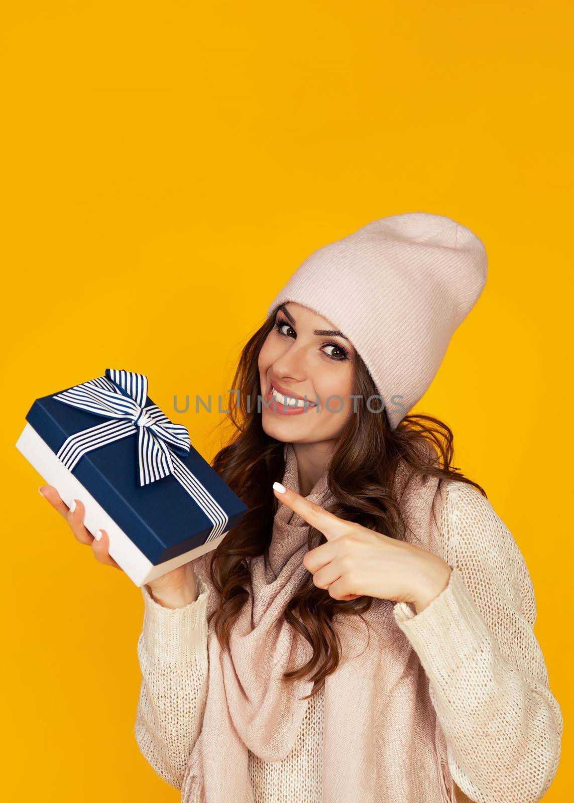 Happy young woman smiling pointing finger holding blue gift box in hand. A gift to your beloved man. A girl in a sweater looks at the camera and points to a gift. Christmas New Year holiday concept