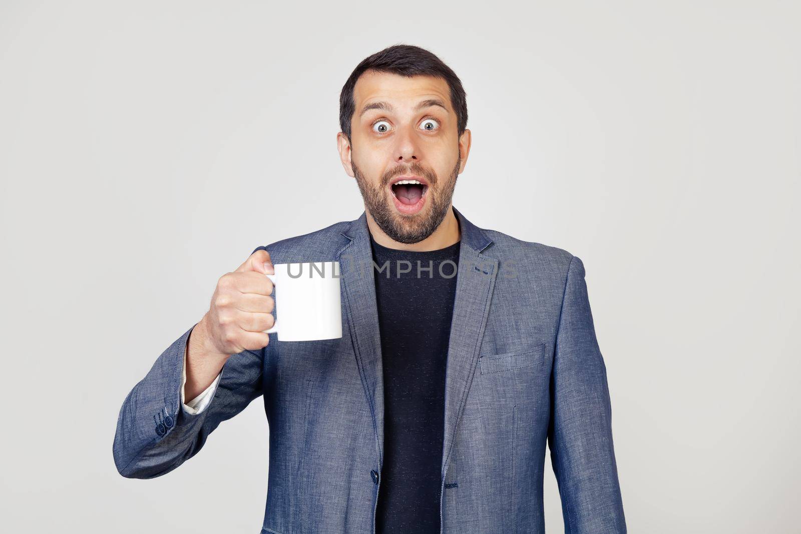 Young businessman man with a beard in a jacket, holding a cup of coffee, scared in shock with a surprised face, scared and excited with an expression of fear. Portrait of a man on a gray background by ViShark