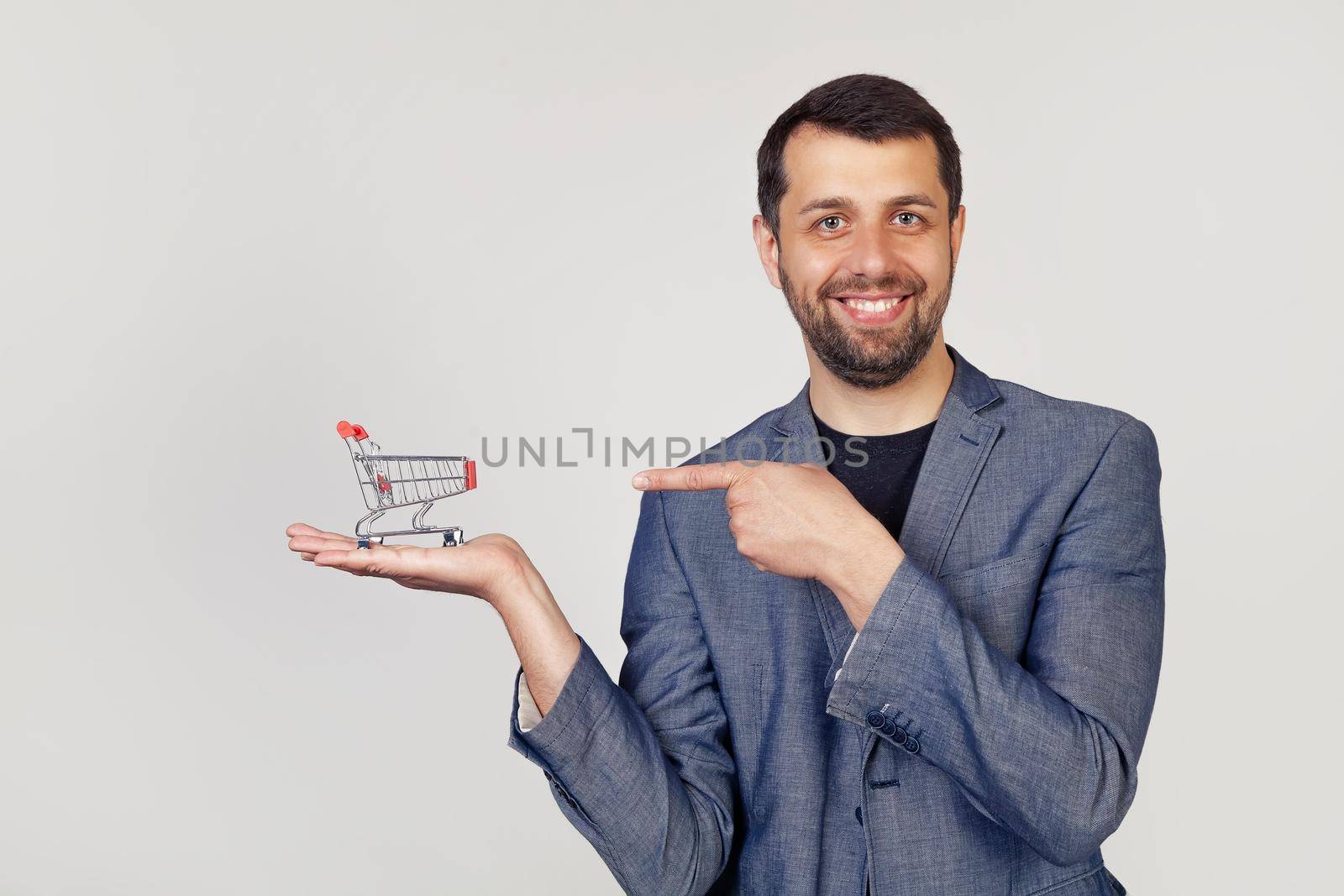 Young businessman man with a beard in a jacket looks at the camera and points to the shopping cart on his hand, showing with his hand and pointing with his finger