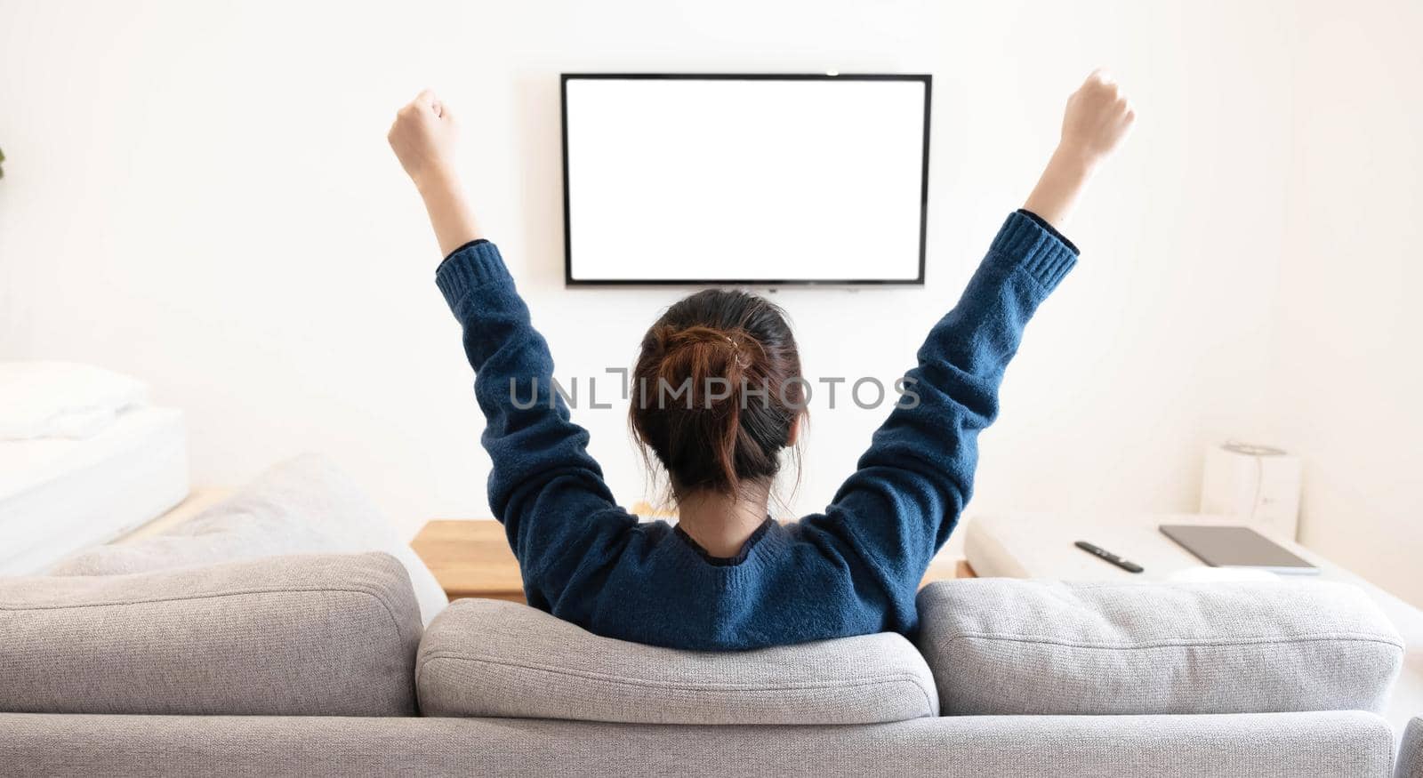 behind of asian young woman watching white screen TV with victory on sofa at living room home at home.