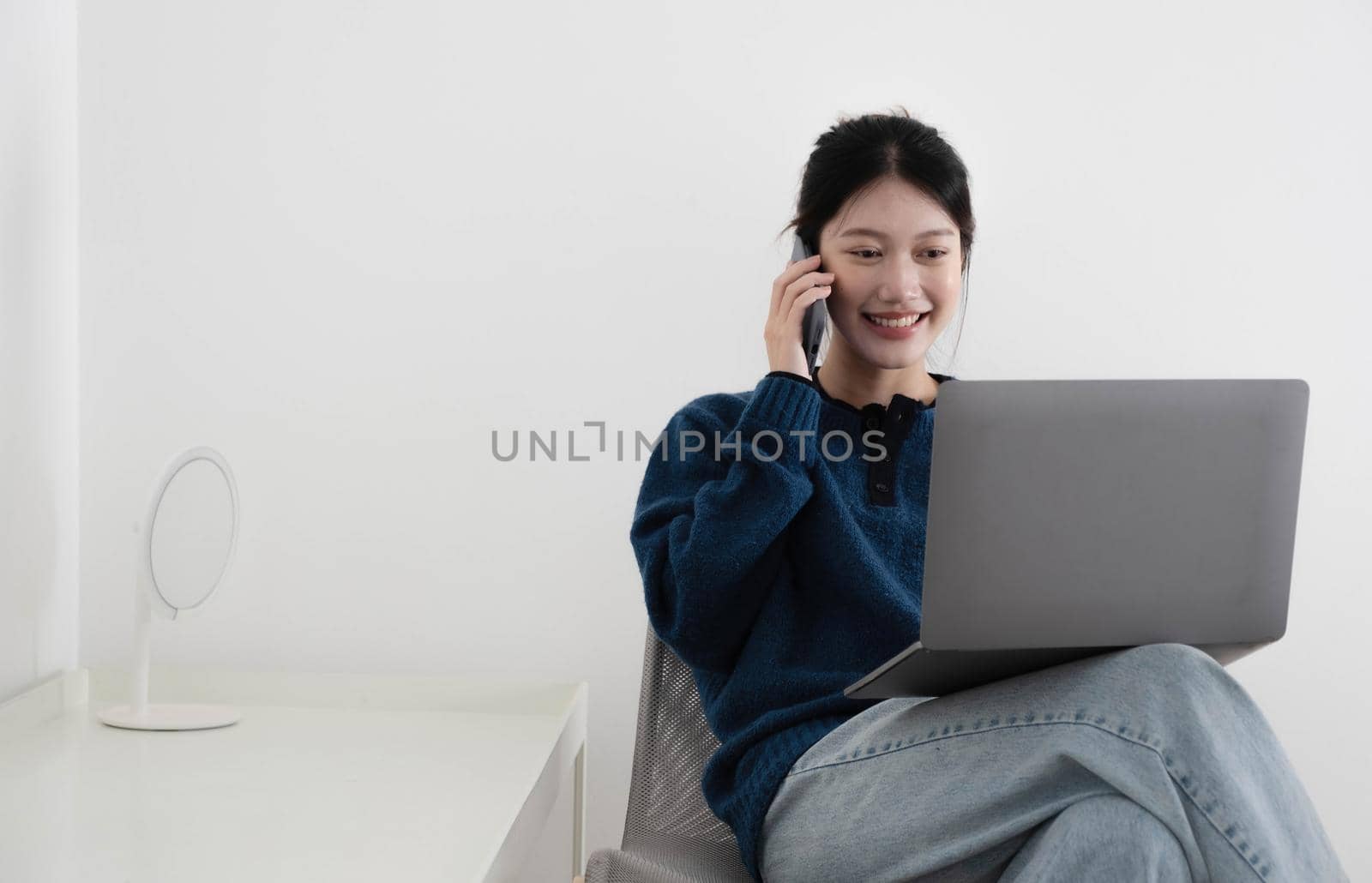 portrait of a happy casual asian woman holding laptop computer and hold smartphone while sitting on a chair over white background.