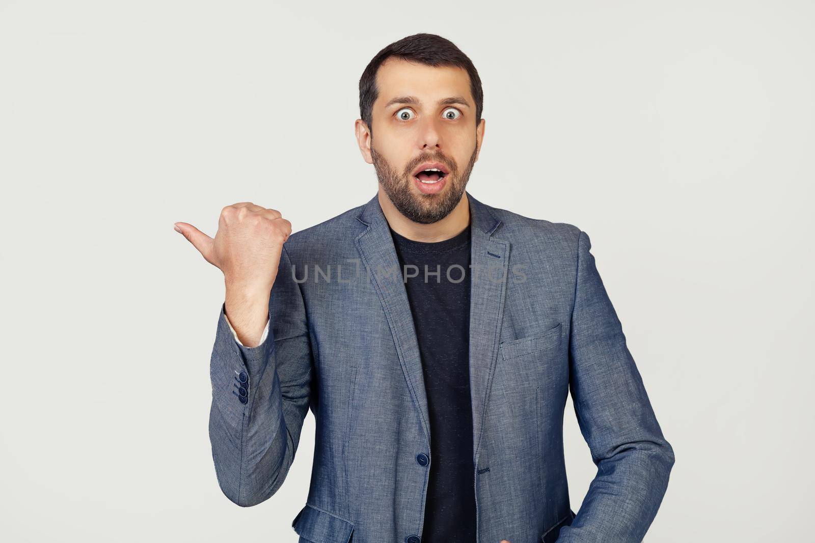 Young businessman man with a beard in a jacket, surprised, pointing finger to the side, with open mouth amazed expression. Portrait of a man on a gray background.