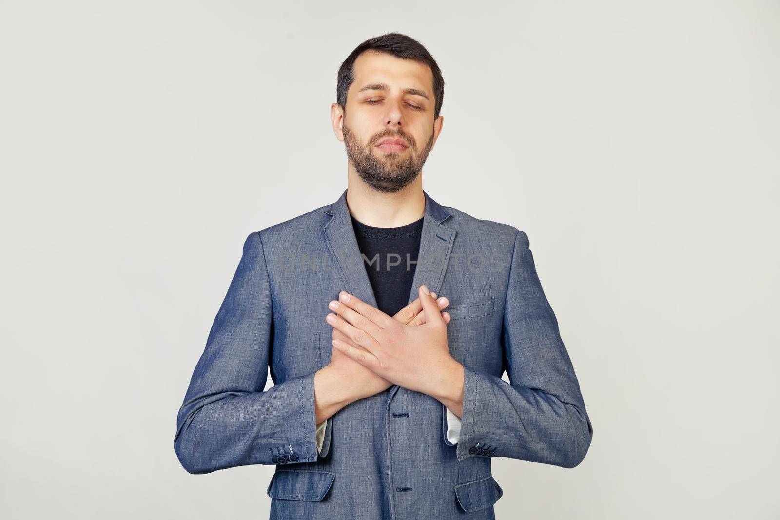 Young businessman with a smiling man with a beard in a jacket, with hands on his chest with closed eyes and a grateful gesture on his face. Health concept. Portrait of a man on a gray background by ViShark