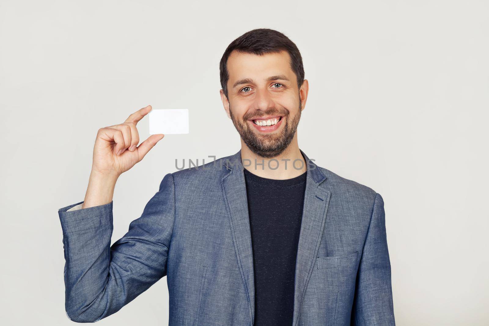 Young businessman man with a beard in a jacket showing a blank sheet of paper. The man smiles and holds in his hand shows a business card. Portrait of a man on a gray background.