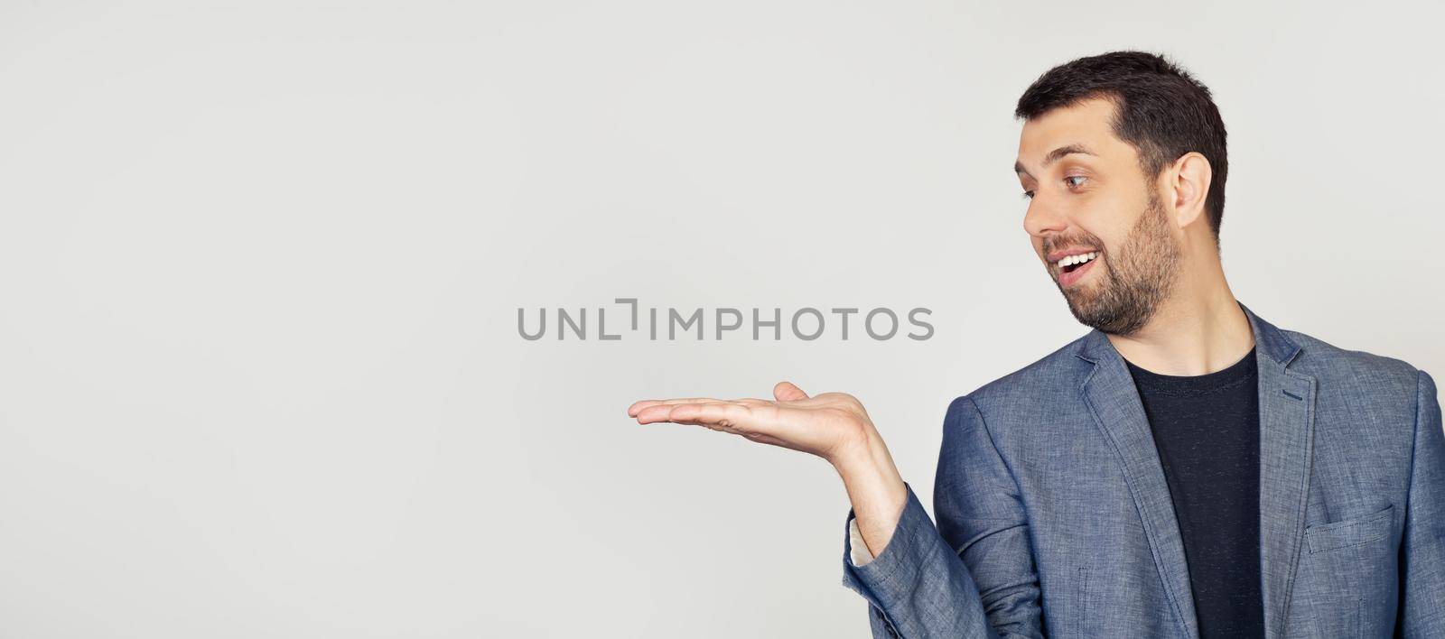 Young man with a beard in a white t-shirt smiling, cheerful presenting and pointing with his palm, looking at his hand. Stands on isolated yellow background