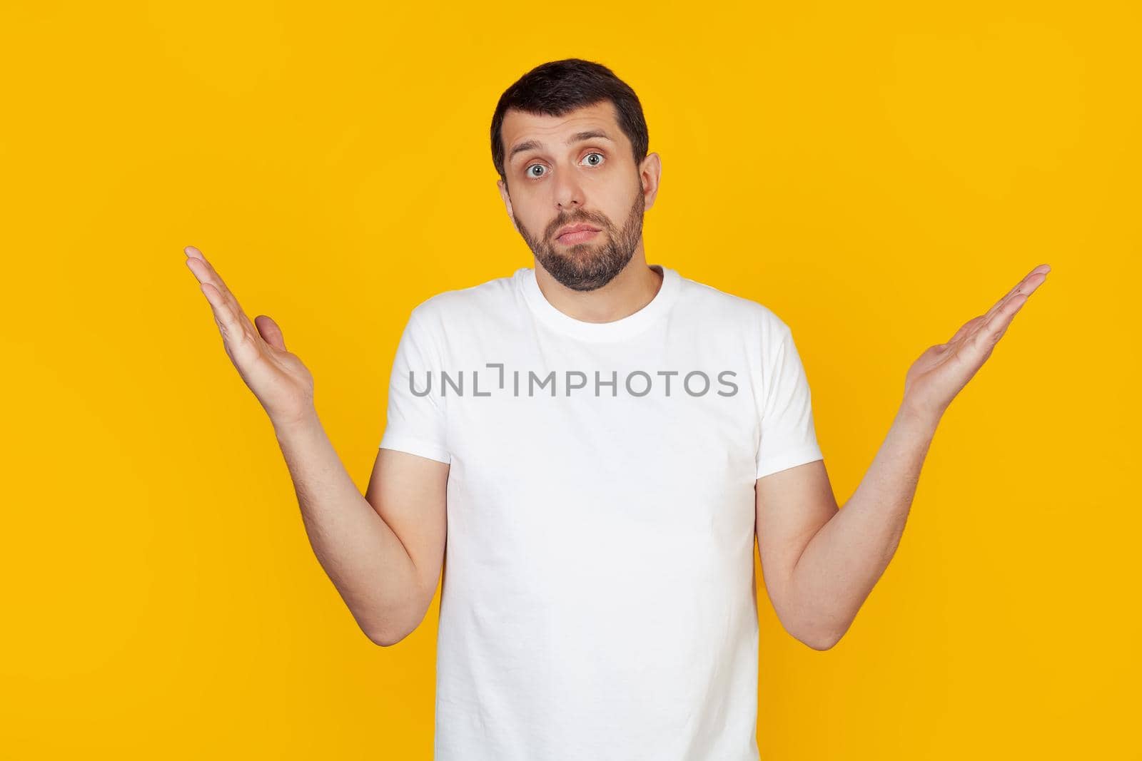 A young man with a beard in a white t-shirt is an ignorant and confused expression with raised arms and hands. Doubt concept. Stands on isolated yellow background.