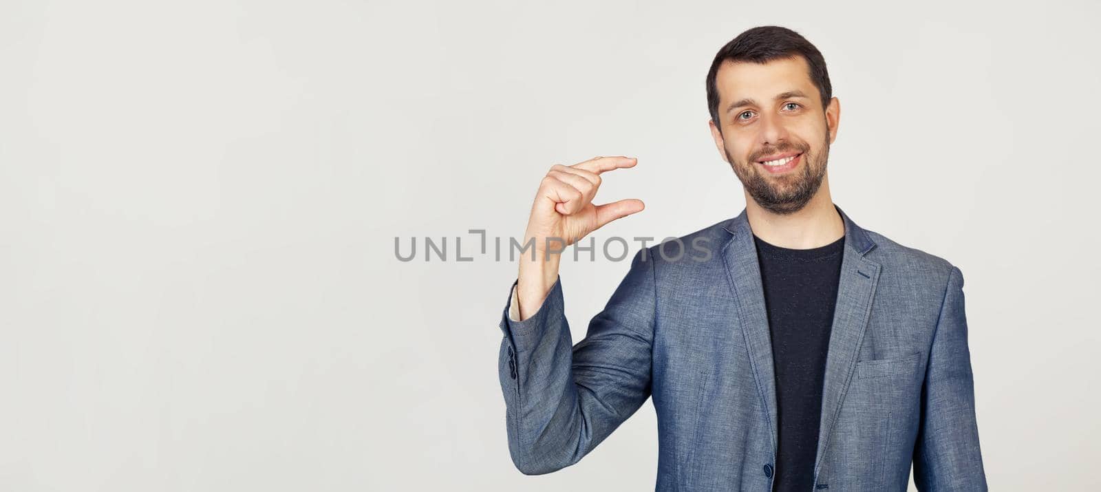 Young businessman man with a beard in a jacket, smiling and confidently gesturing with his hand, making a small size sign with fingers looking and the camera. Measurement concept. gray background.