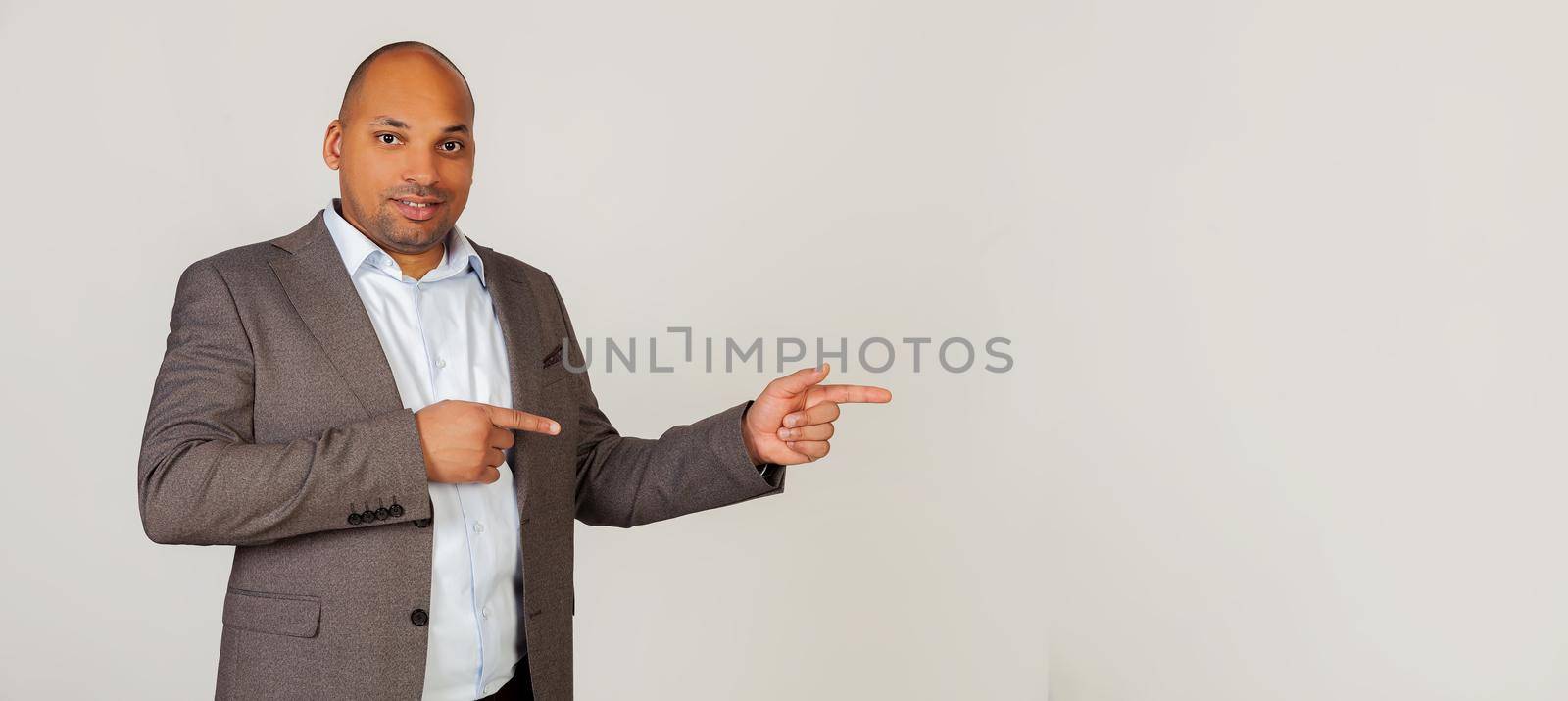 Young african american guy businessman looking into the camera smiling happily and pointing to the side with his index fingers on both hands. Copy space. Standing on a gray background