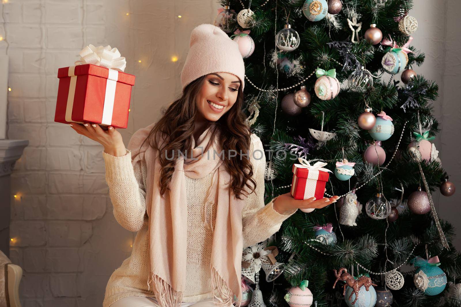 Smiling and happy young woman in winter clothes sitting comfortably on the floor near the Christmas tree opens a New Year's present. The girl enjoys Christmas gifts, meets the New Year and Christmas