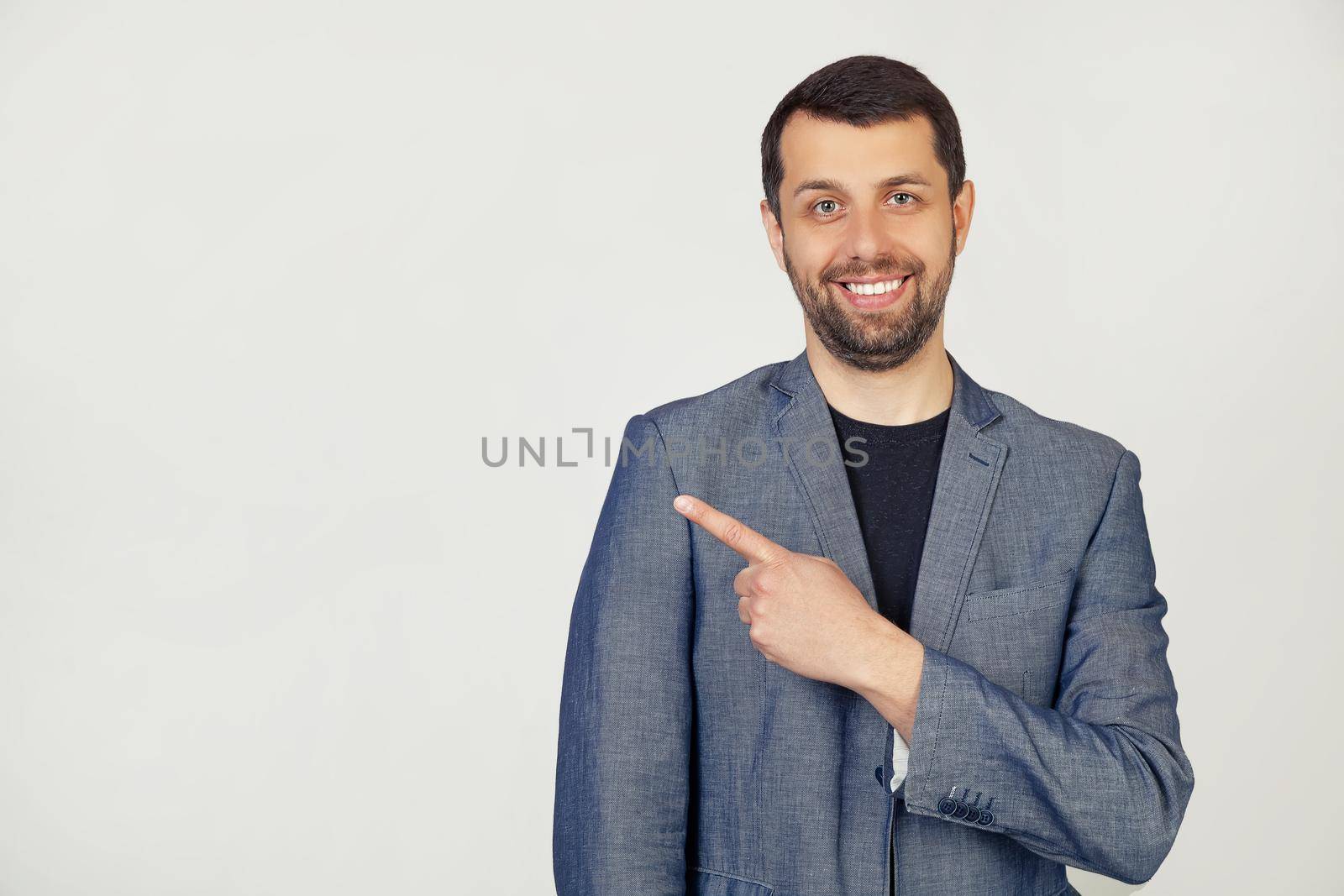 Young businessman man with a beard in a jacket, cheerful, with a smile on his face, pointing with his hand and finger to the side with a happy and natural expression. Portrait of a man on a gray background.