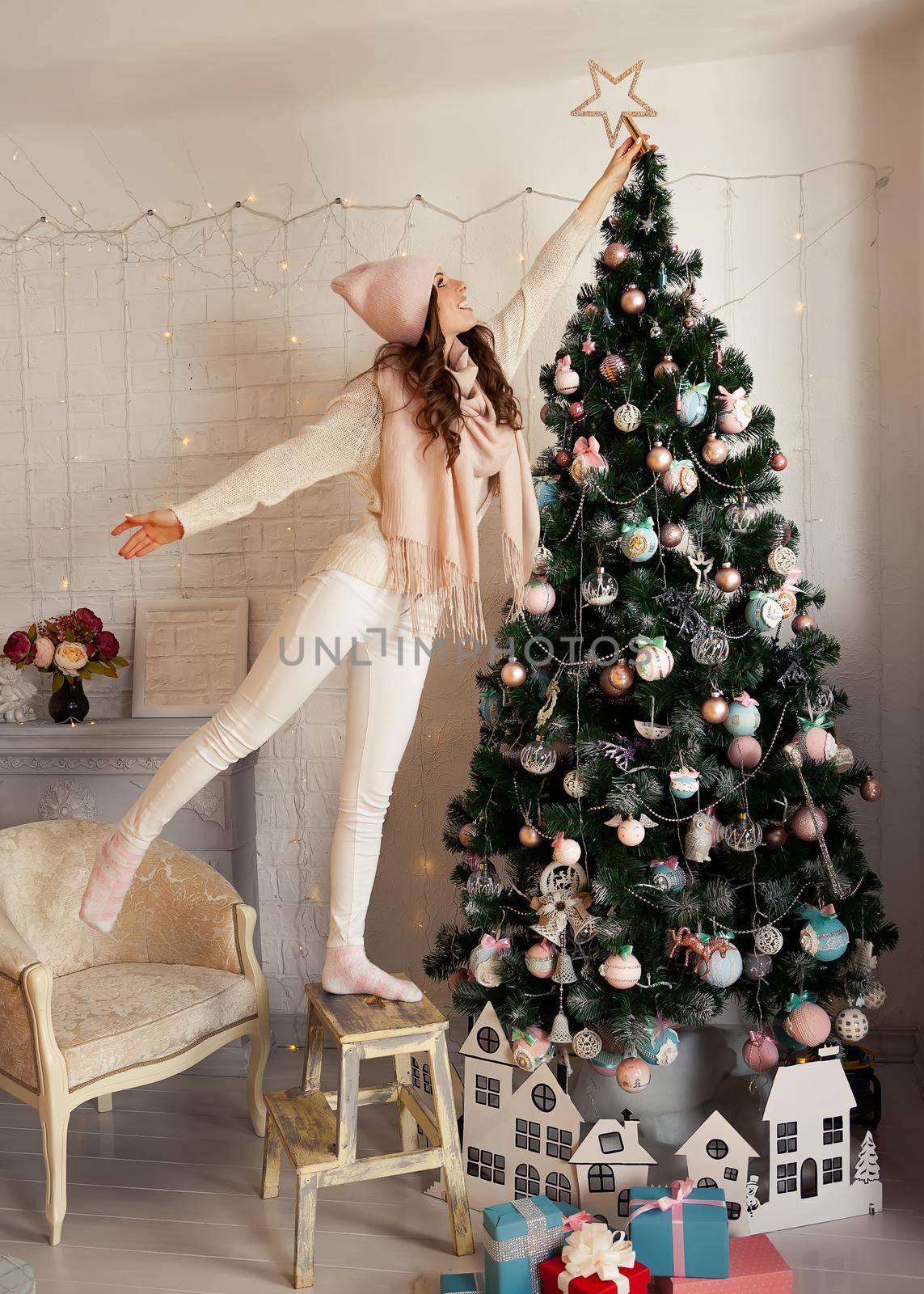 Happy young woman decorates the Christmas tree. A girl in a sweater sets a star on top of a Christmas tree. New year and christmas holiday concept