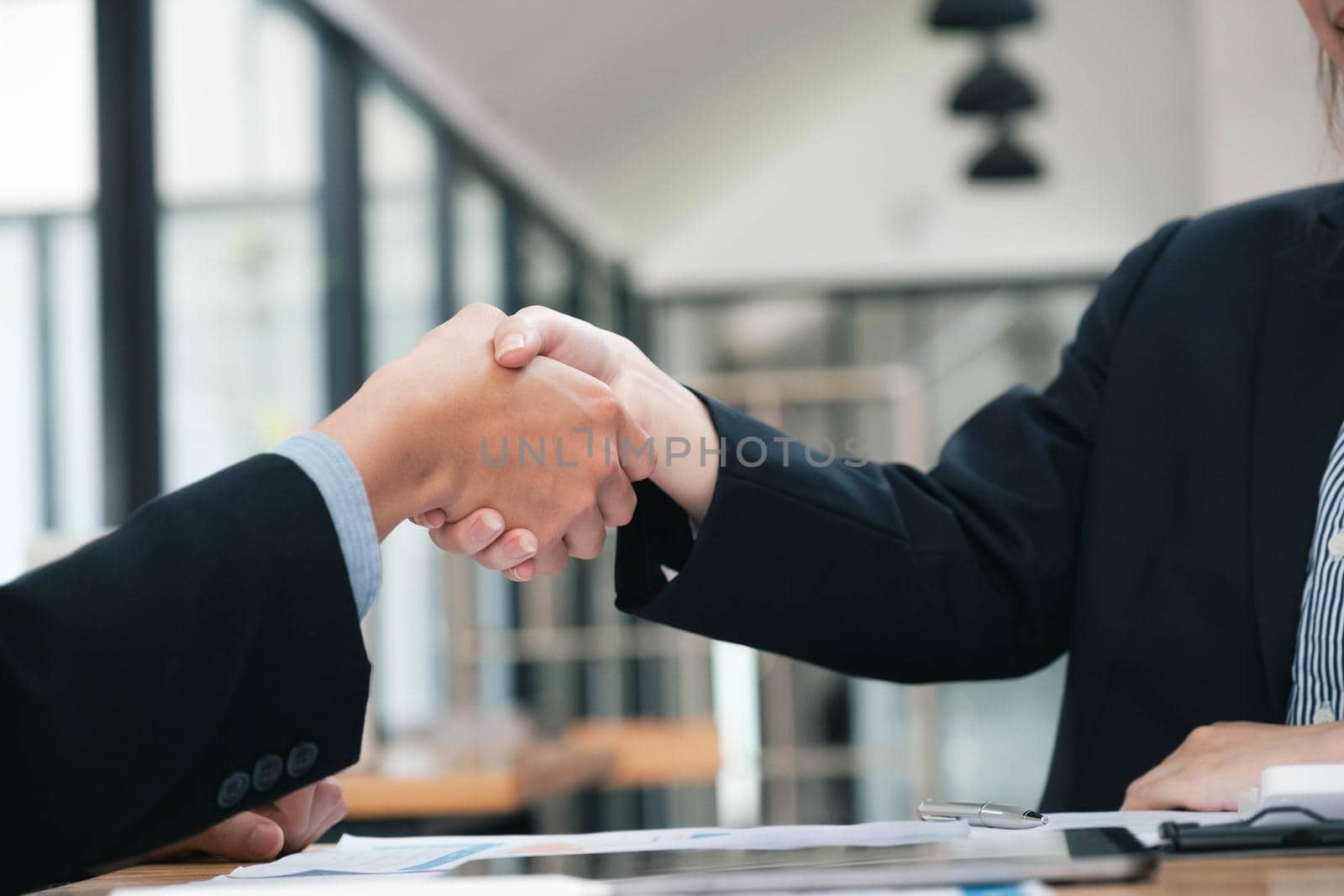 Unknown businesspeople are shaking their hands after signing a contract, while standing together in a sunny modern office, close-up. Business communication, handshake, and marketing concept.