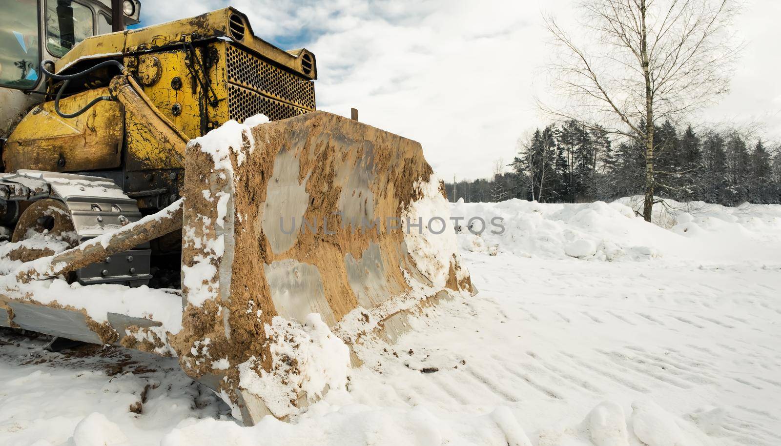 Yellow crawler tractor bulldozer with a bucket works in winter clearing the road from snow. Frosty day with bright sun and beautiful blue sky in the background. Yellow bulldozer in winter