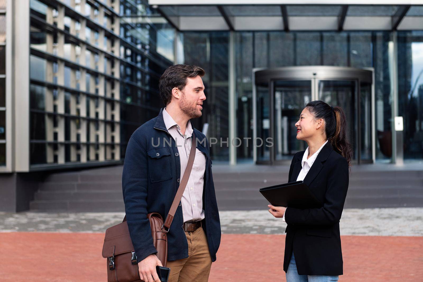 caucasian man and asiatic woman talking about work in front of the entrance to an office building, concept of business and lifestyle, copy space for text