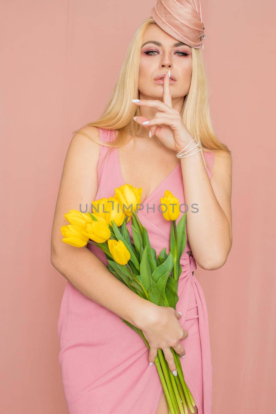 Adult blonde woman in pink dress posing over rosy background. Spring-summer clothing. She is holding bouquet of yellow tulips in her hands. Presents, surprise