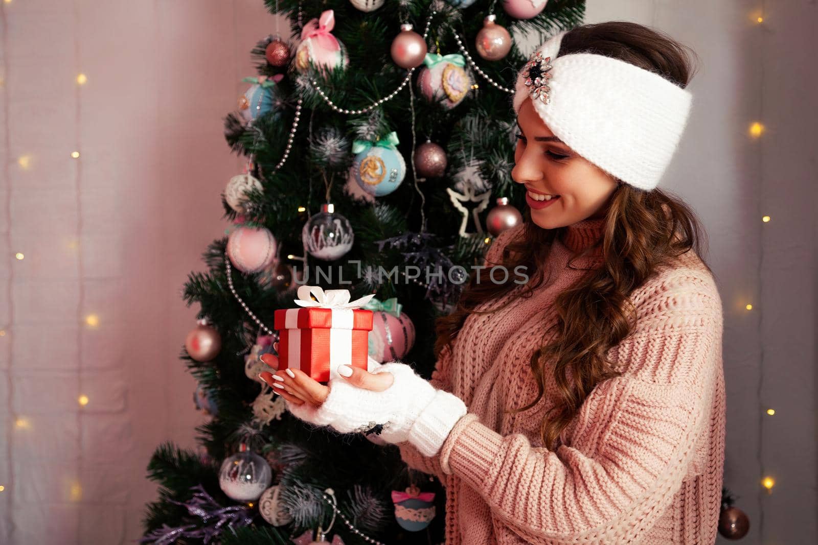 Happy woman holding a gift box on the background of a Christmas tree. Portrait of a happy smiling girl looking at a delivered wrapped box near a decorated fir tree.