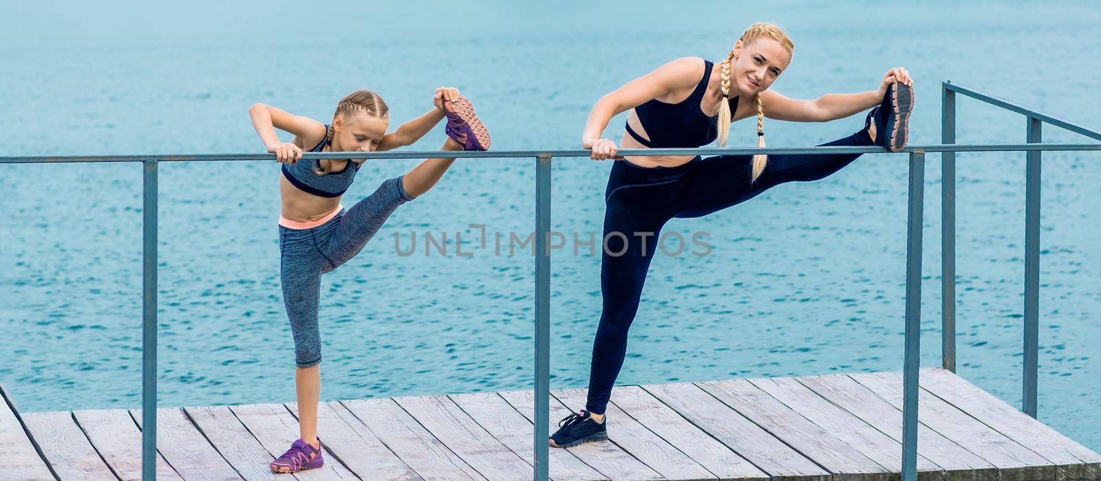 Sportive happy mother is doing workout with little daughter on the shore of the lake.