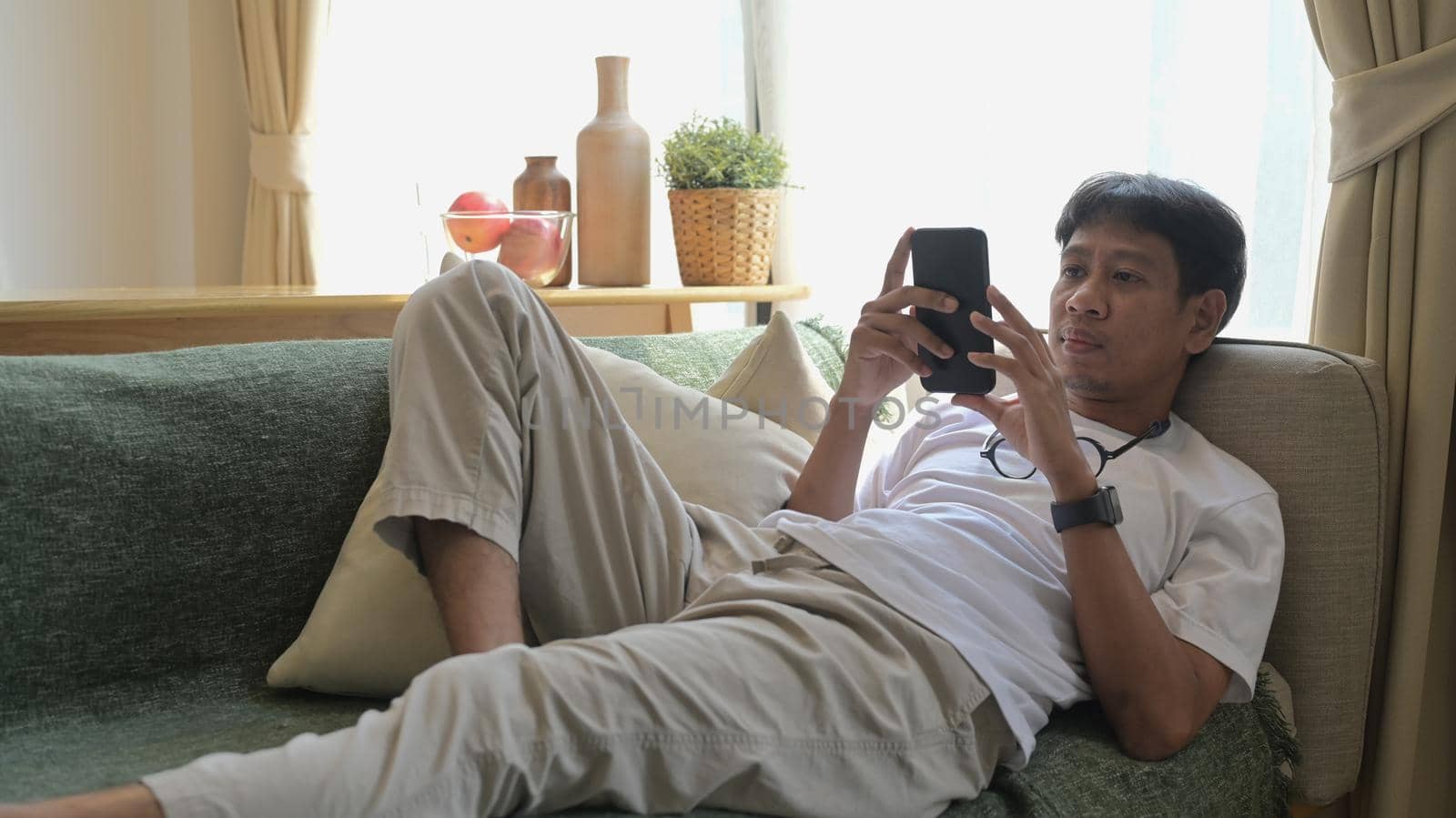 Relaxed man lying on couch in bright living room and using mobile phone for for communicating in social network.