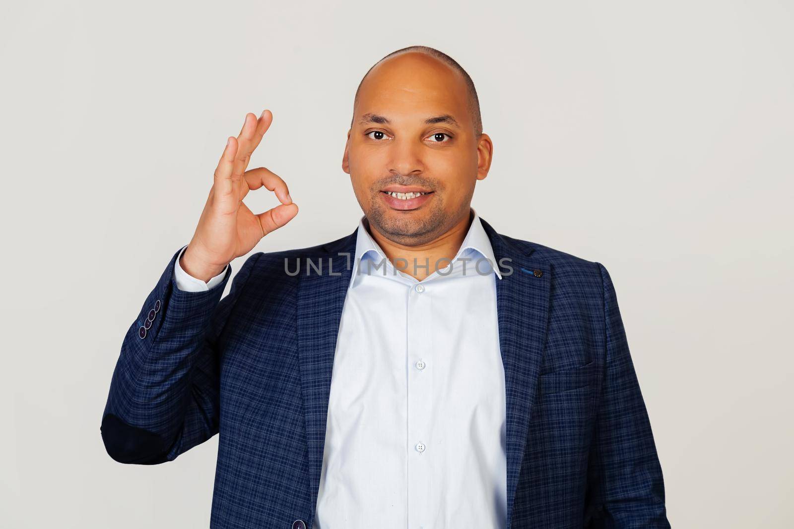Portrait of a young African American guy businessman, smiling positively, making okay sign with hand and fingers. Lucky expression. Standing on a gray background. by ViShark