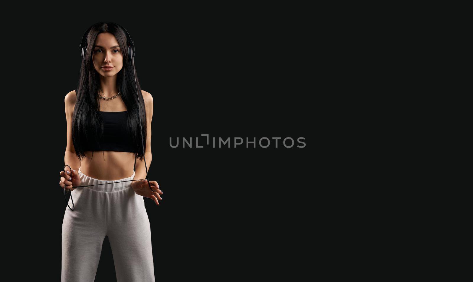 Front view of brunette girl with long hair listening to music in earphones. Pretty slim female wearing black top and white trousers standing, looking ar camera. Concept of youth.