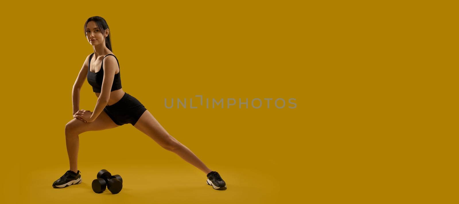 Side view of pretty slim young female working out indoors. Girl with ponytail wearing black top and shorts, stretching, making step, dumbbells on floor. Concept of youth.