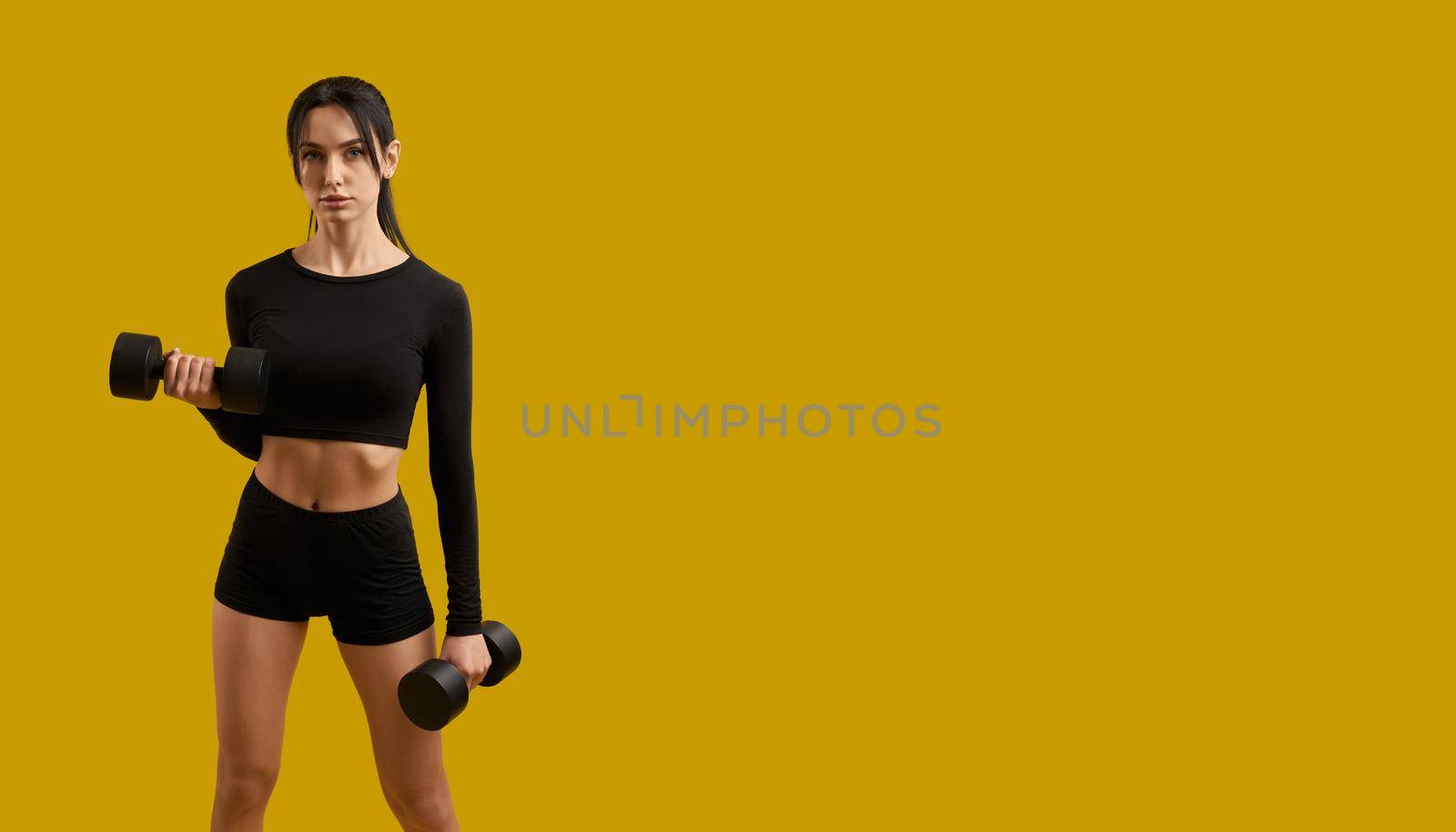 Front view of pretty slim girl in sportswear working out, training, exercising. Brunette female with ponytail standing, raising dumbbells, looking at camera. Concept of youth.