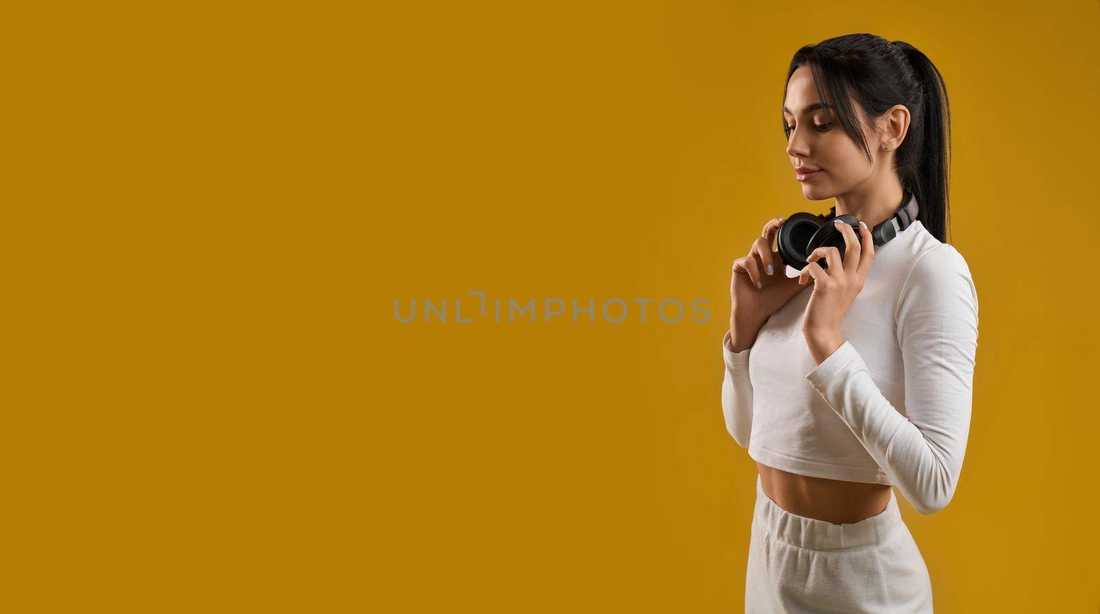 Side view of brunette girl with ponytail standing, looking down. Pretty slim young female wearing white top and trousers, holding earphones on neck. Concept of modern life.