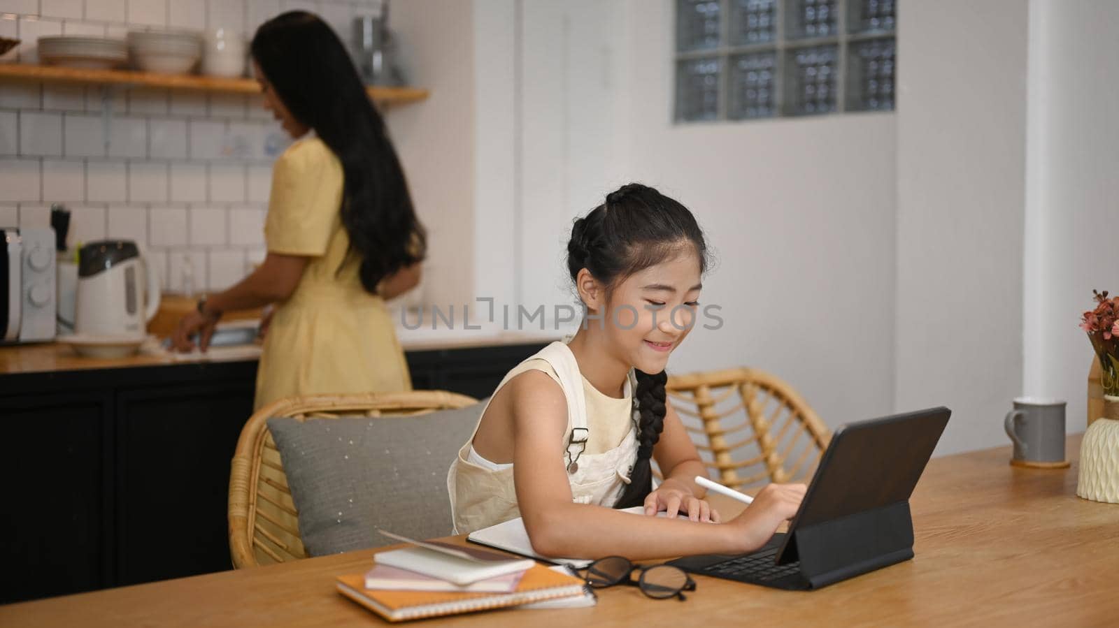 Asian girl using laptop during online class with tutor or teacher while sitting in home kitchen and mother in background by prathanchorruangsak