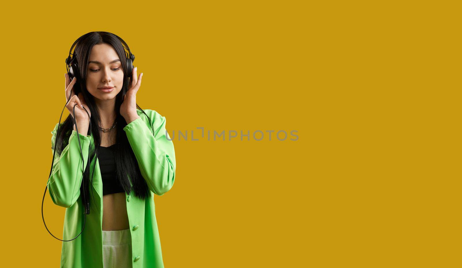 Front view of girl wearng green suit, standing, listening to music. Pretty brunette woman with long hair enjoying music with closed eyes, smiling. Concept of modern life.