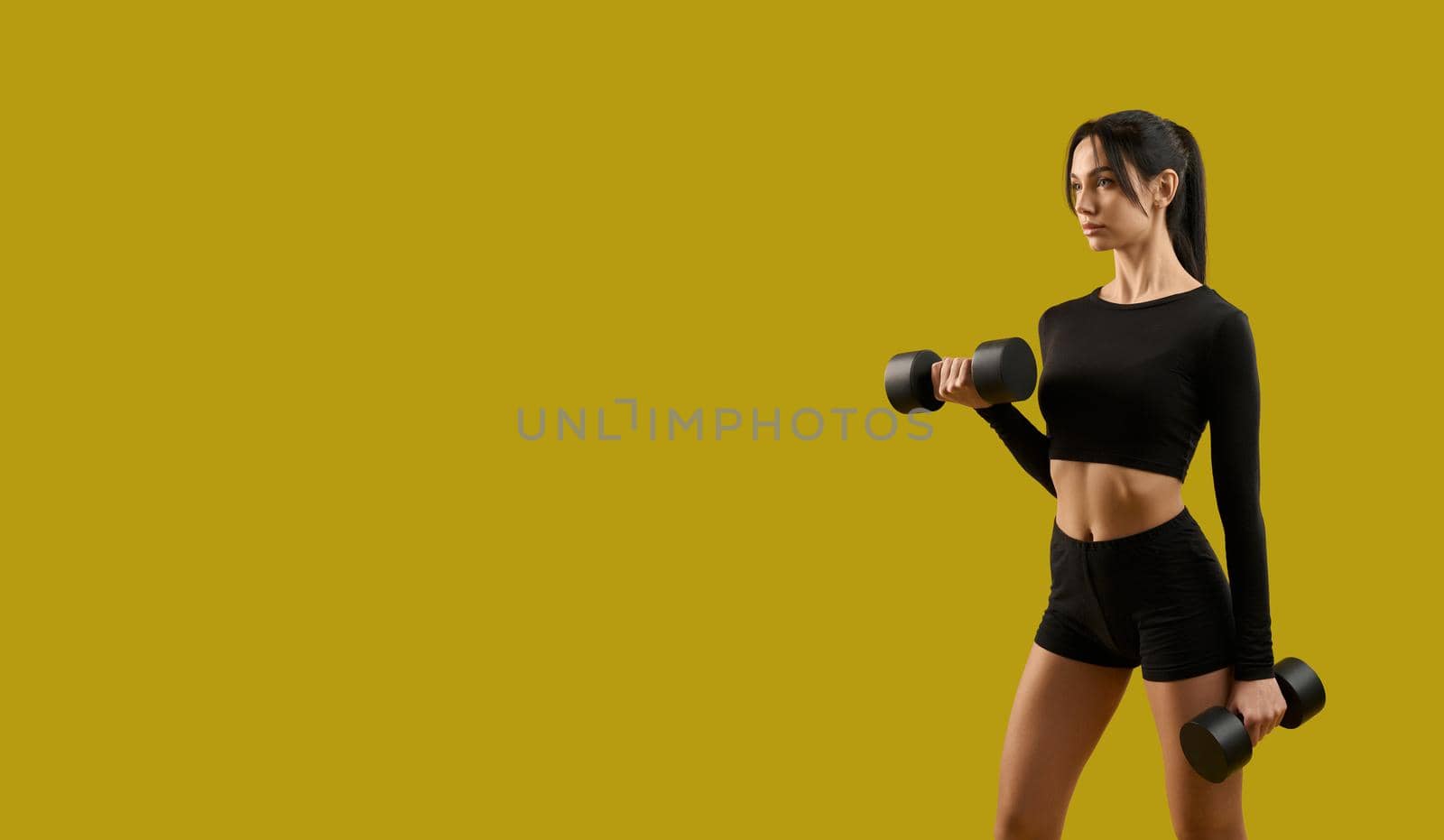 Side view of pretty slim girl standing, raising dumbbells. Brunette young woman making step, working out, stretching, looking forward. Isolated on yellow studio background.