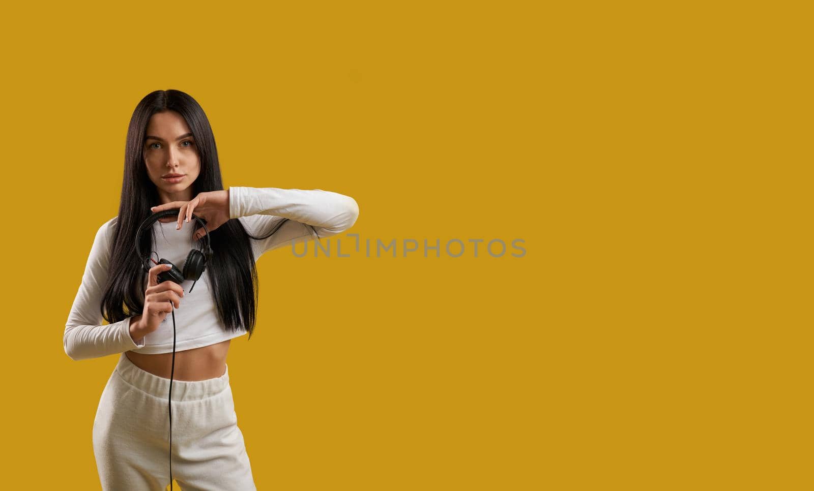 Front view of brunette girl with long hair standing, holding hands. Pretty slim young female wearing white sports suit, looking at camera, serious. Concept of modern life.