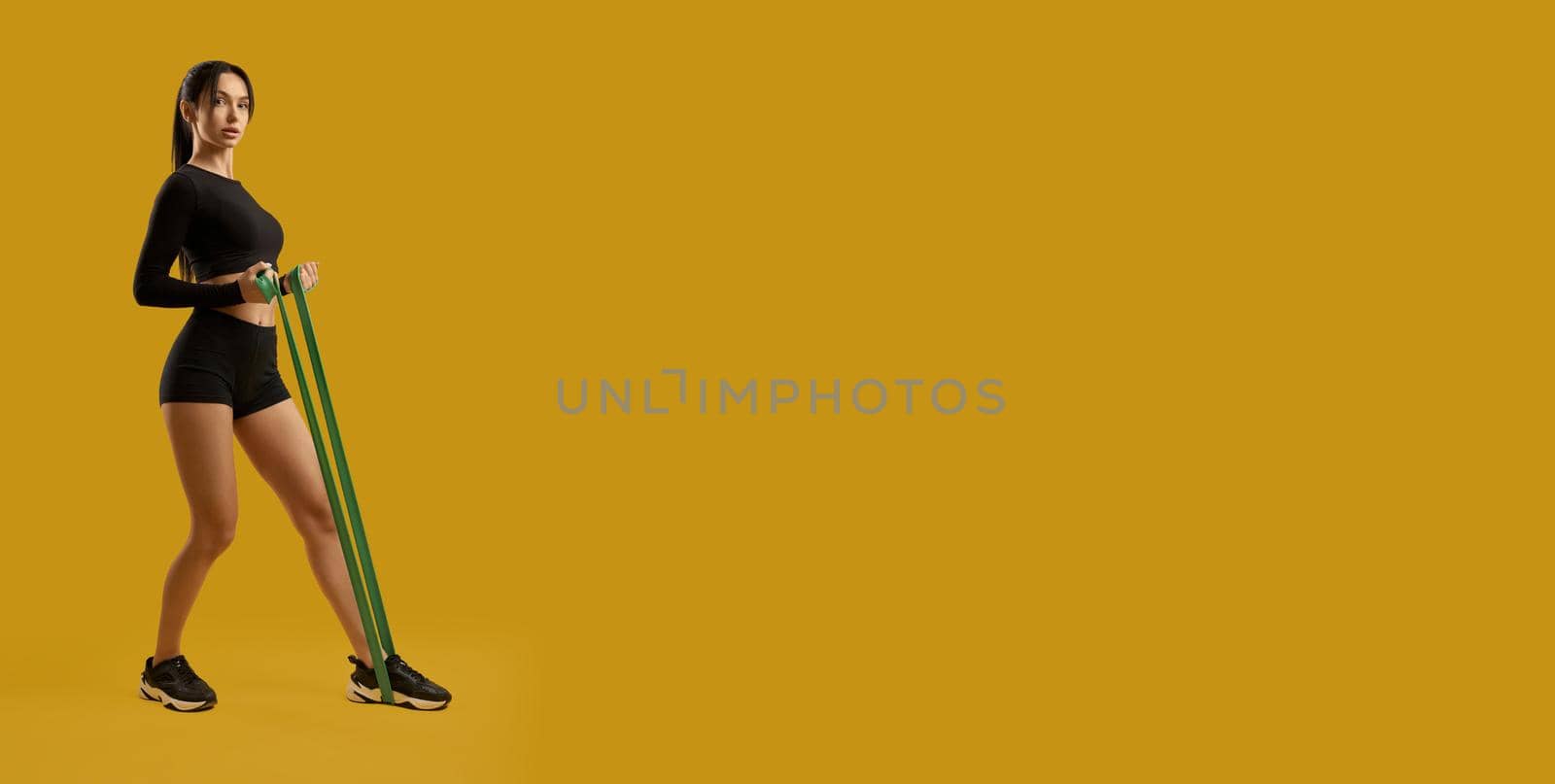 Side view of brunette sporty female exercising, training with rubber bands. Pretty slim girl standing, making step, stretching, serious. Isolated on yellow studio background.