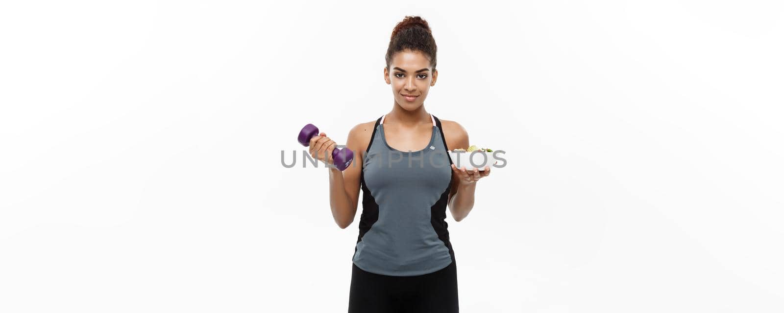 Healthy and Fitness concept - Beautiful sporty African American on diet holding dumbbell and fresh salad on hands. Isolated on white studio background