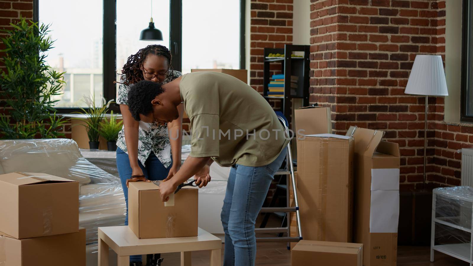African american couple wrapping cardboard boxes with sticky tape to move in new apartment flat. Packing up furniture and home belongings in packages with adhesive roller for relocation.