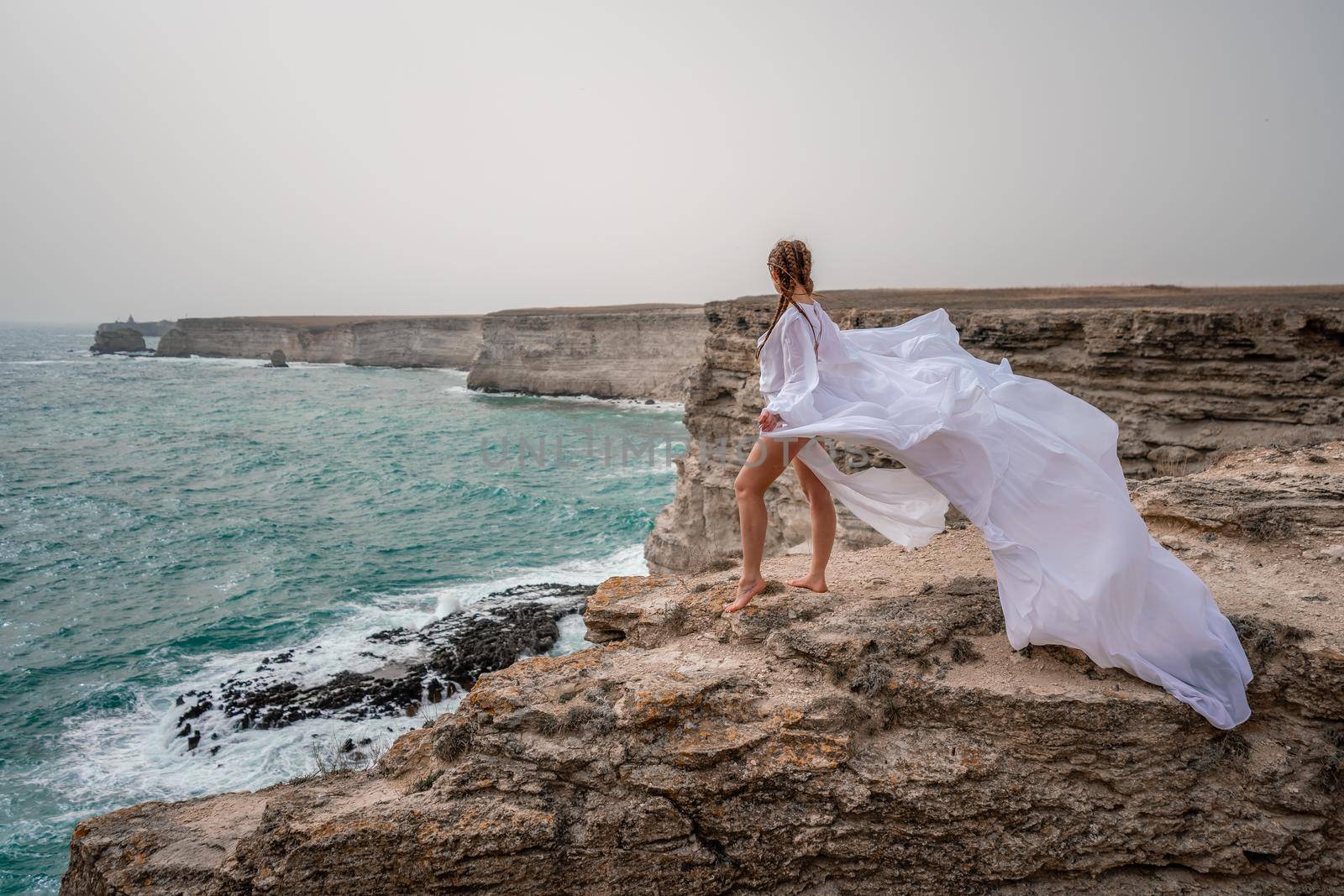 Happy freedom woman on the beach enjoying and posing in white dress over the sea. View of a girl in a fluttering white dress in the wind. Holidays, holidays at sea