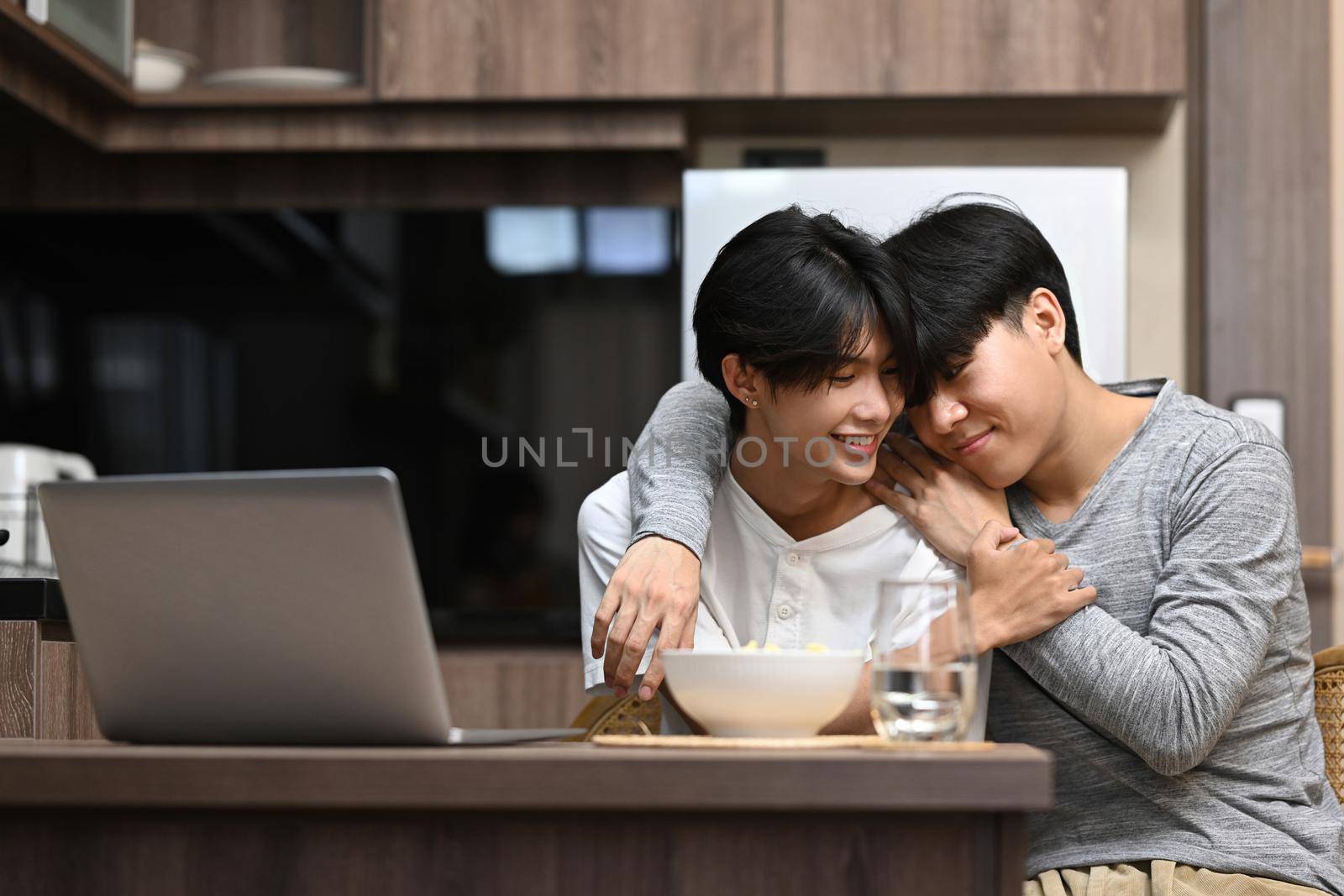 Sweet homosexual couple having a happy morning conversation and embraced while sitting in the kitchen by prathanchorruangsak