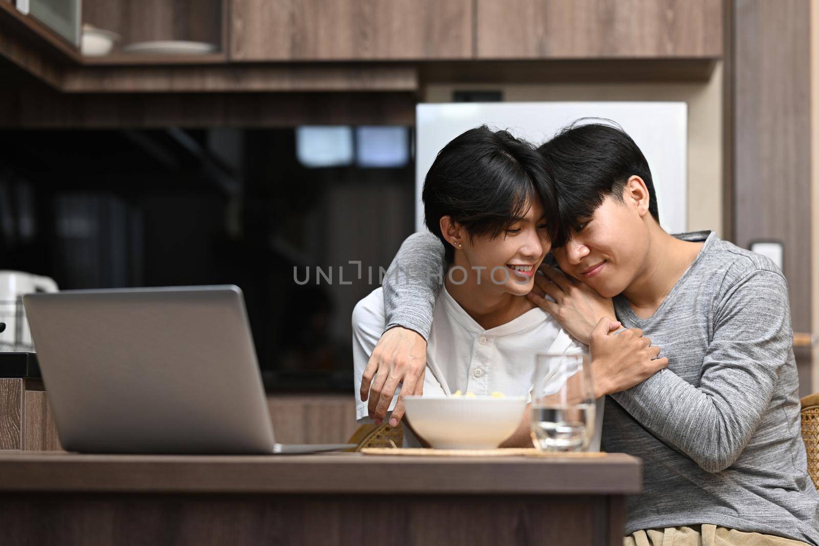 Sweet homosexual couple having a happy morning conversation and embraced while sitting in the kitchen.