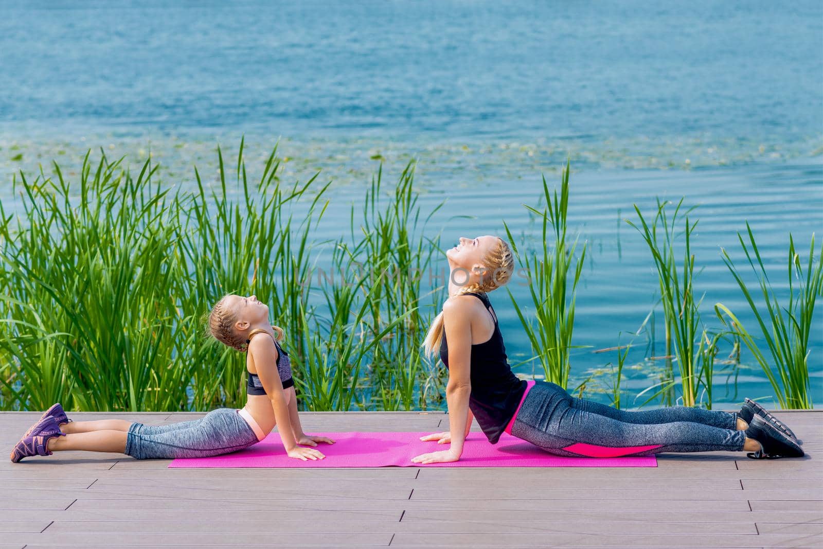 Little girt and woman are doing exercises on the grass at the shore of the lake.