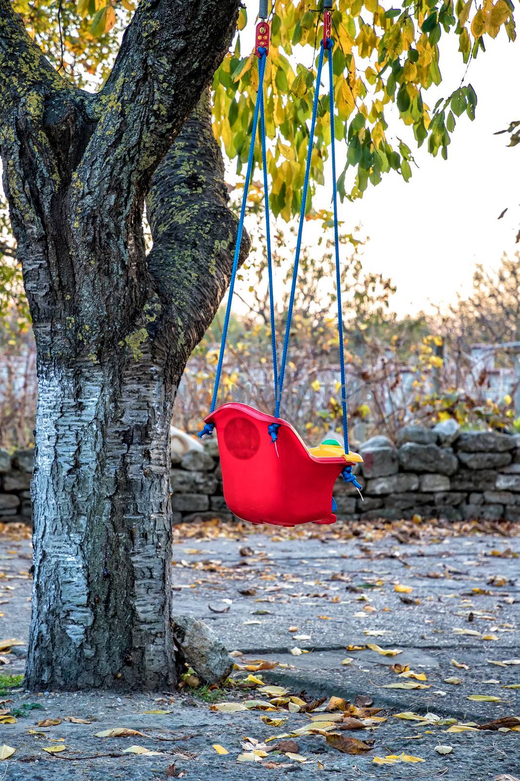 Empty red plastic swing at the tree in the garden, fun outdoor activity for kids