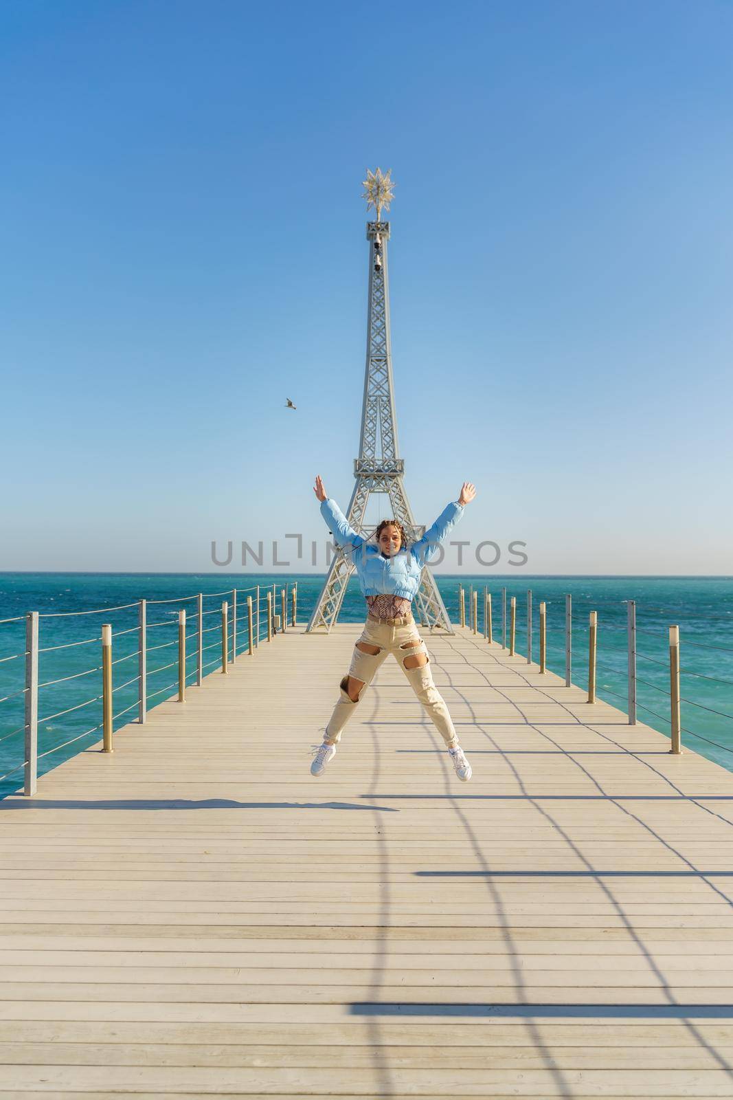 Large model of the Eiffel Tower on the beach. A woman walks along the pier towards the tower, wearing a blue jacket and white jeans. by Matiunina