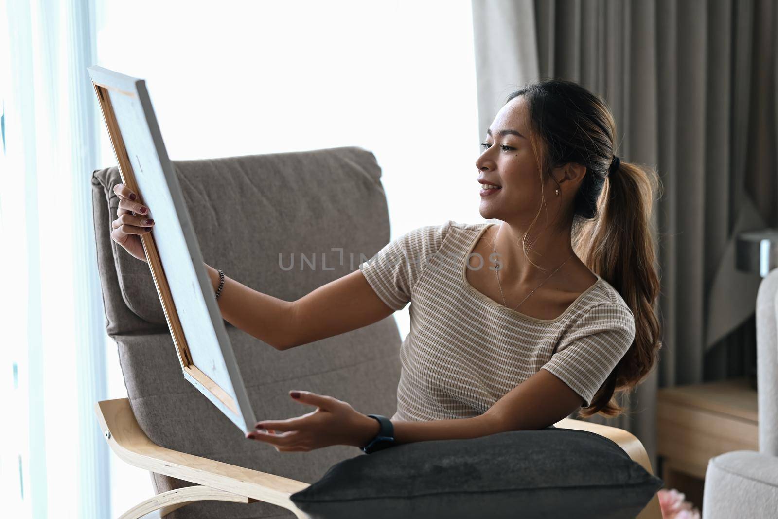 Happy young woman sitting on armchair in living room and looking at her artwork with admiration.