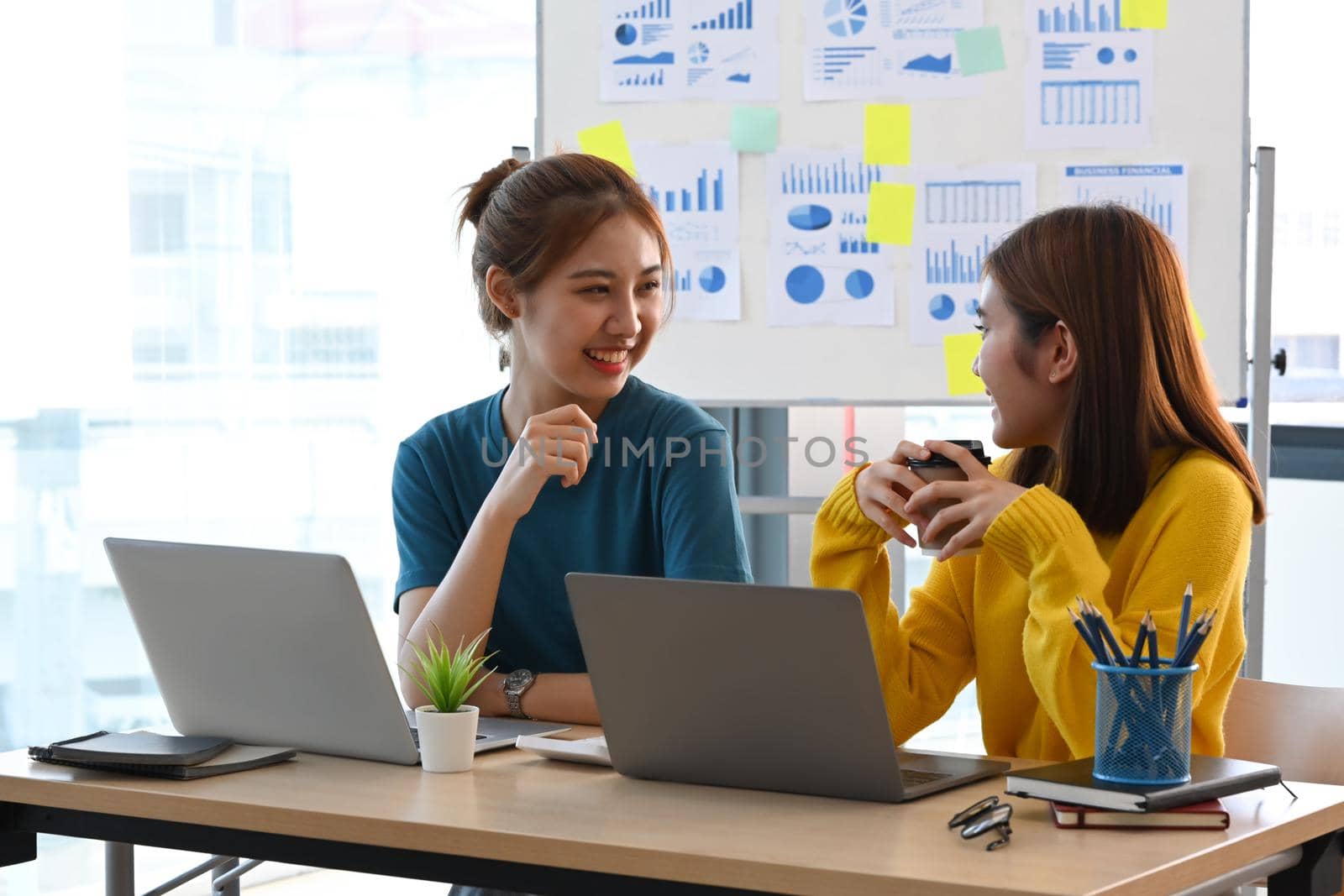 Two cheerful creative women talking each other while working together at modern office. by prathanchorruangsak