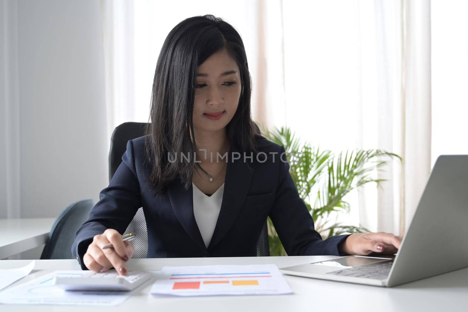 Asian business woman using calculator and working with laptop at office desk.
