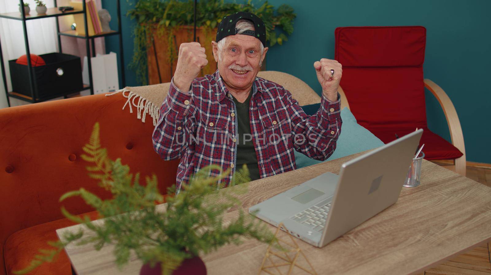 Senior old grandfather man in stylish cap playing computer video games on laptop computer, wins, rises hands, celebrate, smiles happily. Gamer, having fun, rest. Virtual reality. Modern technologies